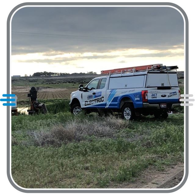 A white and blue utility truck with Magic Valley Electric on the side is parked on grassy terrain near cultivated fields under a cloudy sky. A person stands nearby working, with farmland and hills in the background.