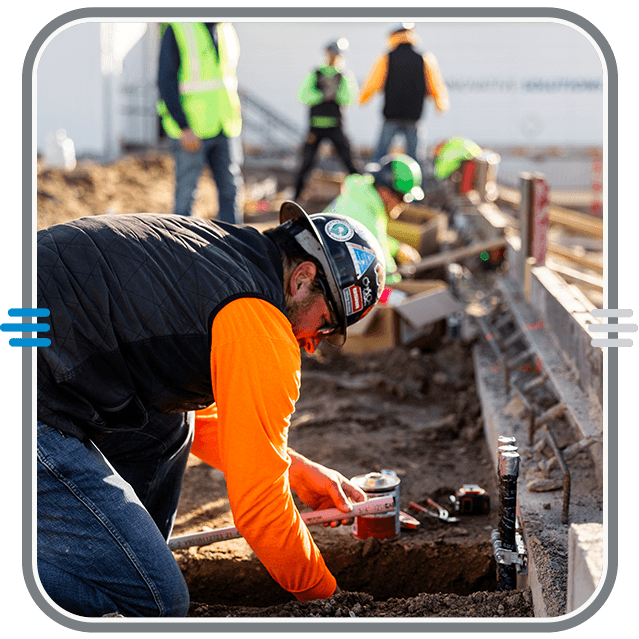 Construction workers wearing safety gear work on a building site, with one worker in the foreground using tools to work in a dirt trench, while others work in the background.