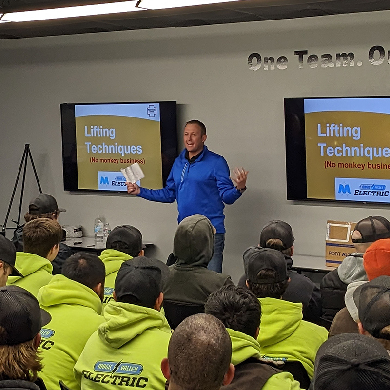 Team meeting with man presenting at front of the group. There are TVs behind him with "Lifting Techniques (No Monkey Business)" on the televisions.