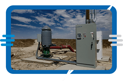 Outdoor industrial equipment setup on a dirt ground, including a control panel, pipes, and a cylindrical tank, under a partly cloudy sky with barren land in the background.