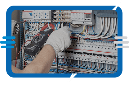 A person wearing a white glove uses a multimeter to test electrical wires inside an open electrical panel filled with various colored cables and circuit breakers.